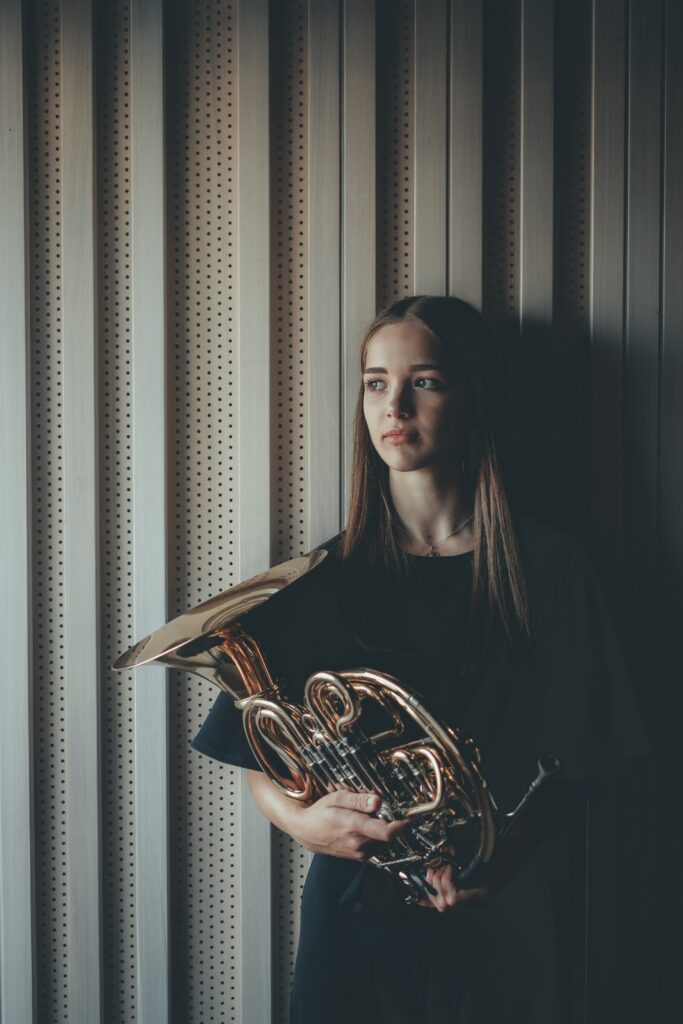 A woman hold a French horn in front of a grey wall