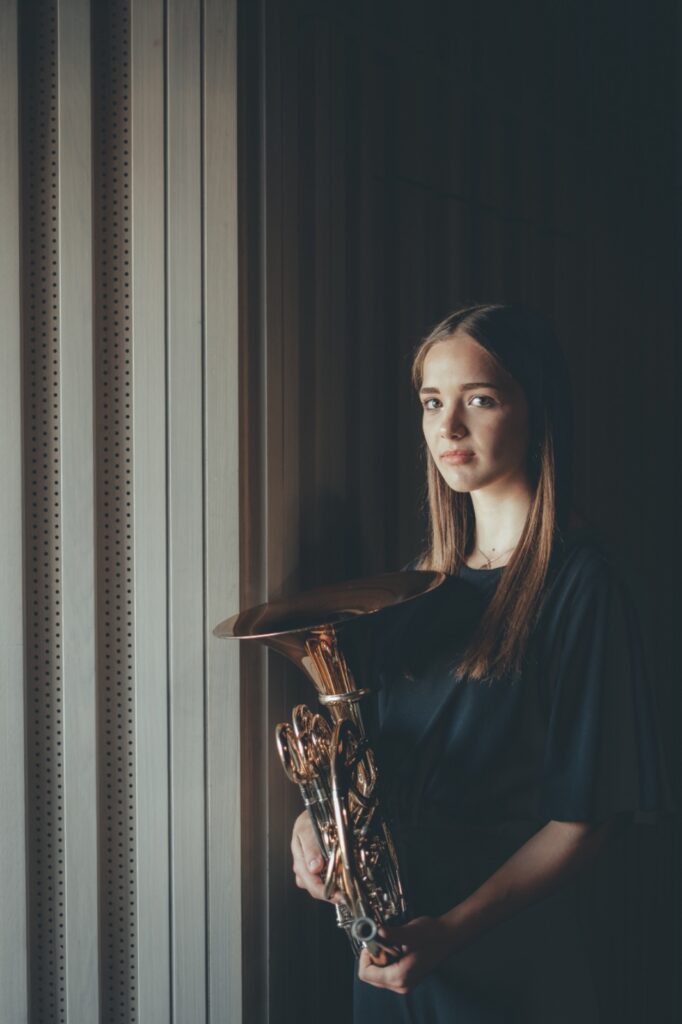A woman holds a French horn, in front of an inside wall